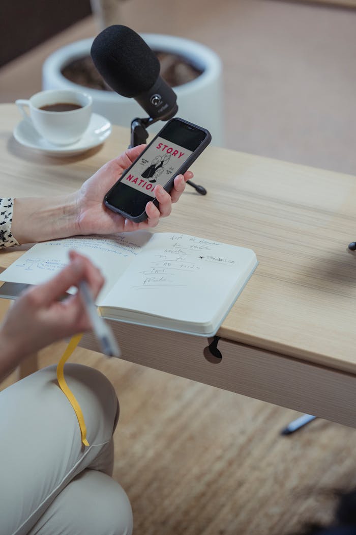 A person recording a podcast using a smartphone and taking notes at a desk.